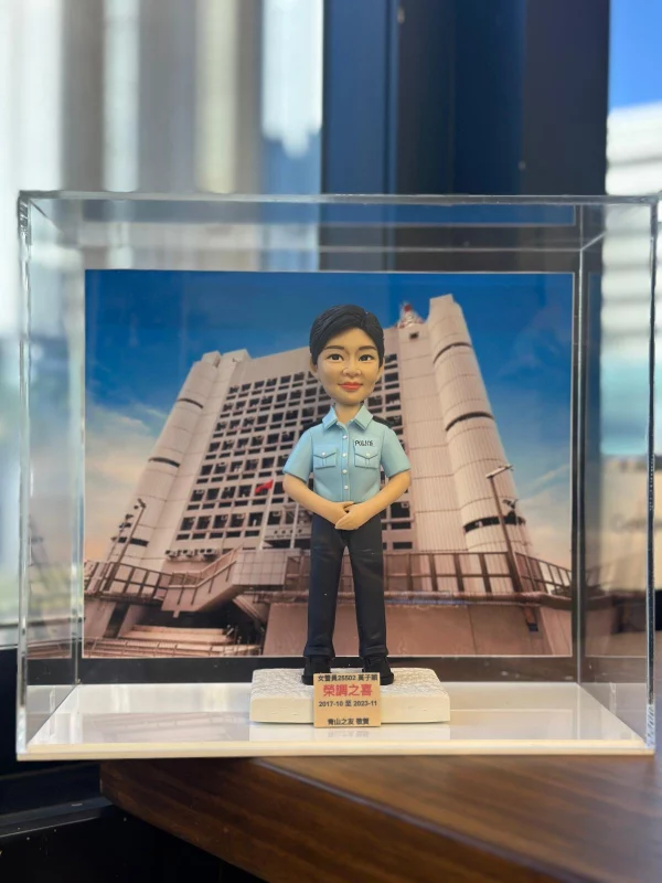 一款為香港女警員訂製的榮調紀念人像公仔，公仔身穿警察制服，背景為荔枝角收押所，由其同事為祝賀她調職而敬贈。