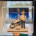 一個透明展示盒內的警察主題人像公仔場景。公仔頭身比例可愛化，短髮、戴深藍色貝雷/軟帽，眉眼以手作刻線描出微笑神情；身穿淺藍短袖制服襯衫配深藍長褲與黑色皮鞋，襯衫有口袋、摺線與鈕扣刻紋，左肩掛有對講機肩咪與線材，肩章與領角描邊清晰。人物雙手於腹前合掌呈立正站姿。腳下是淡灰石紋地台，左側擺放一台白色警車模型（車頂與車身可見POLICE字樣、藍紅條紋與車燈結構）。地台前緣貼有金屬色名牌，上印「榮升之喜」等字樣。背板為「New Territories South Regional Police Headquarters｜新界南總區警察總部」主視覺海報，含部門徽章與大樓插畫。展示盒置於窗邊拍攝，可見室外街景與高樓的淡化倒影；前景有一隻手從下方托住盒子。