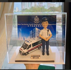 一個透明展示盒內的警察主題人像公仔場景。公仔頭身比例可愛化，短髮、戴深藍色貝雷/軟帽，眉眼以手作刻線描出微笑神情；身穿淺藍短袖制服襯衫配深藍長褲與黑色皮鞋，襯衫有口袋、摺線與鈕扣刻紋，左肩掛有對講機肩咪與線材，肩章與領角描邊清晰。人物雙手於腹前合掌呈立正站姿。腳下是淡灰石紋地台，左側擺放一台白色警車模型（車頂與車身可見POLICE字樣、藍紅條紋與車燈結構）。地台前緣貼有金屬色名牌，上印「榮升之喜」等字樣。背板為「New Territories South Regional Police Headquarters｜新界南總區警察總部」主視覺海報，含部門徽章與大樓插畫。展示盒置於窗邊拍攝，可見室外街景與高樓的淡化倒影；前景有一隻手從下方托住盒子。
