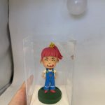 一款客製化的動漫人物人像公仔，一個粉紅頭髮、缺了門牙的淘氣女孩被放置在透明展示盒中，是充滿創意的生日禮物。