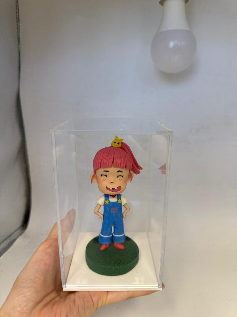 一款客製化的動漫人物人像公仔，一個粉紅頭髮、缺了門牙的淘氣女孩被放置在透明展示盒中，是充滿創意的生日禮物。