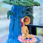 一款超現實主義的動漫人物人像公仔，一個喇叭頭的男子在藍色大樹下的蓮花上冥想，是充滿藝術感的獨特生日禮物。