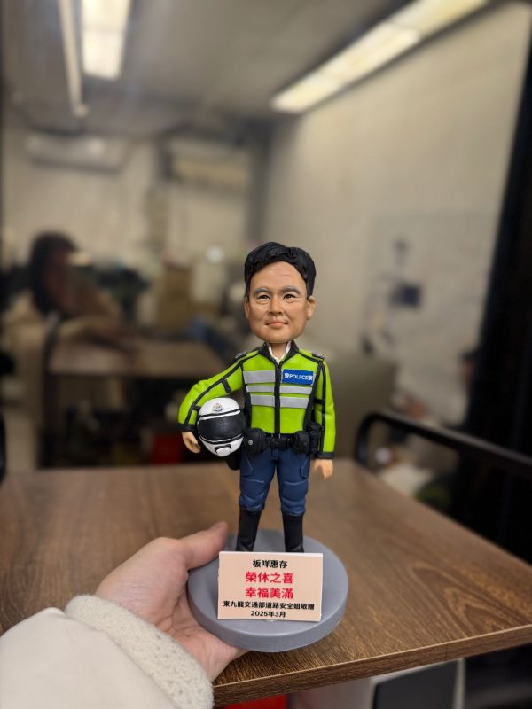 一款為香港交通部警務人員訂製的榮休人像公仔。該警員身穿螢光綠色交通制服，手持電單車頭盔。底座銘牌刻有「榮休之喜」及贈送單位「東九龍交通部執行及管制組」。
