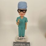 定製醫生人像公仔，角色為外科醫生 Dr. Irene Lo，身穿綠色手術衣與手術帽，配戴護目鏡，手持手術工具，底座寫有「DR. IRENE LO 手術聖手」。此醫護公仔專為外科醫生設計，是獨特的醫療專屬禮物。