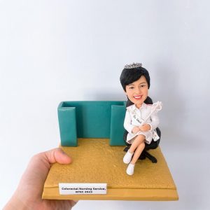 這是一個精緻的手工製作女性護士人像公仔，穿著白色護士制服，頭戴皇冠，手持魔杖。她坐在一張黑色辦公椅上，旁邊有一個綠色的隔板。人像公仔上有一個銘牌，上面寫著“Colorectal Nursing Service, NTEC 2023”。這個公仔展示了一位護士的肖像，應該是代表在結直腸護理領域的醫護專業人士。