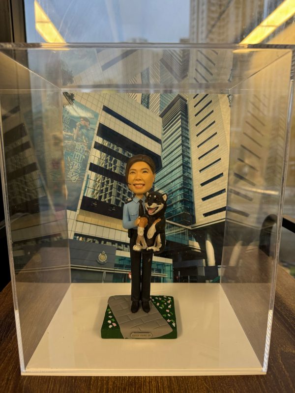 一款祝賀女警務人員晉升的人像公仔禮物，公仔身穿警察制服並懷抱一隻柴犬寵物，背景是警察總部大樓照片。底座銘牌記錄了這次晉升的職級與日期。