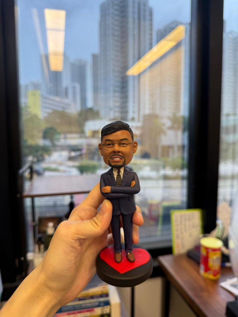 一隻手拿著一個為外國同事訂製的人像公仔,公仔是一位穿西裝、雙臂環抱的男士,站立在一個紅色的心形底座上,適合作為告別禮物。