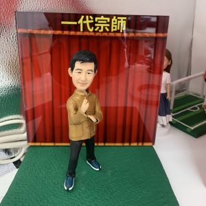 太極公仔，身穿棕色中式唐裝外套、黑色長褲及藍色運動鞋，雙手擺出太極起手式動作，神情專注，背景為紅色幕布，上方有黃色文字「一代宗師」，適合作為武術主題擺設或收藏。