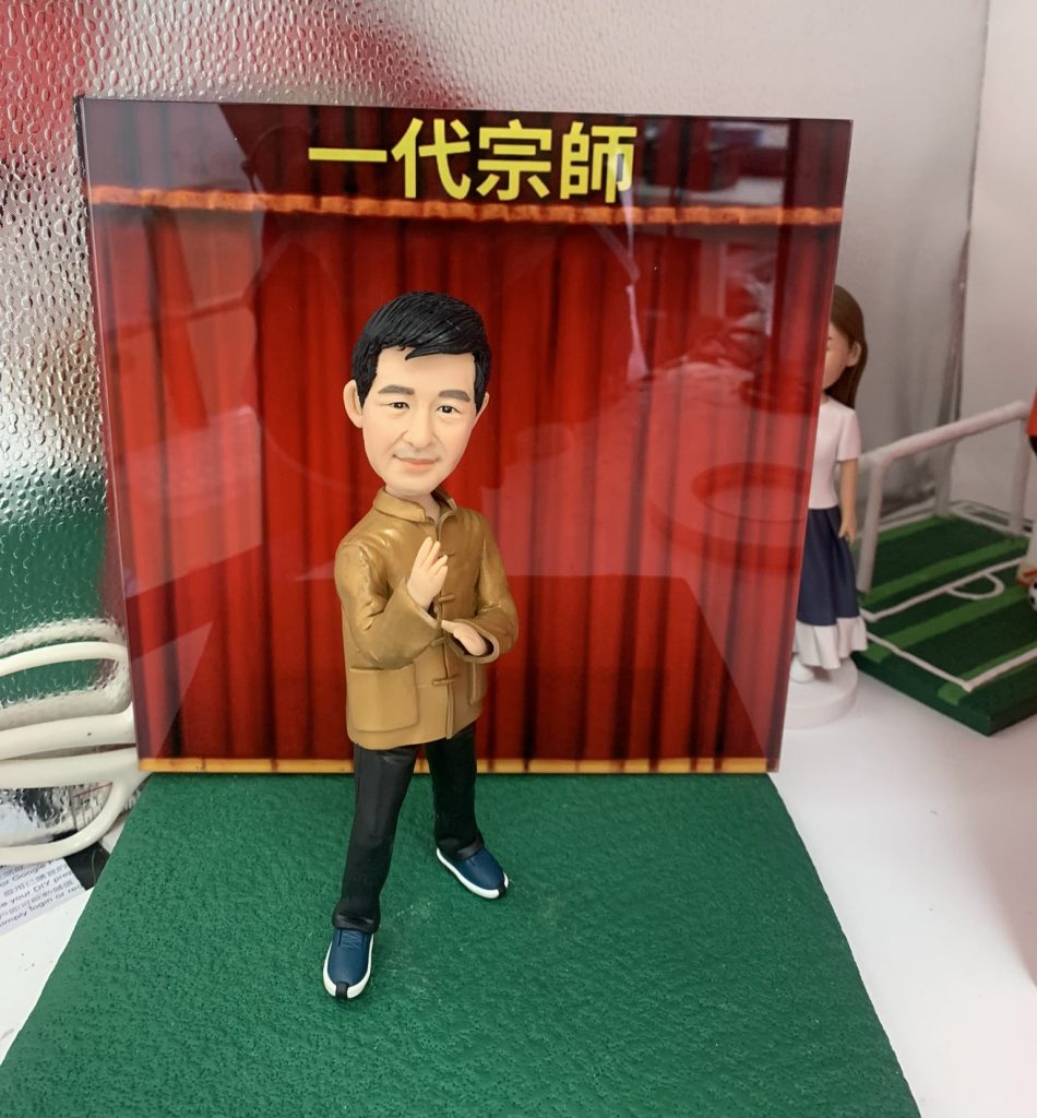 太極公仔，身穿棕色中式唐裝外套、黑色長褲及藍色運動鞋，雙手擺出太極起手式動作，神情專注，背景為紅色幕布，上方有黃色文字「一代宗師」，適合作為武術主題擺設或收藏。