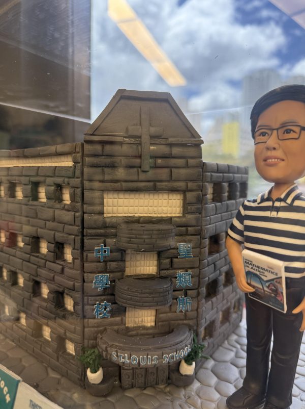一款為聖類斯中學的黎德維校長（Mr. Lai Tak Wai）訂製的校長退休禮物。精緻的人像公仔手持數學書，莊重地站在一座細節豐富的聖類斯中學立體學校模型旁，底座前方陳列著五個社（LILY, ROSE, SHAMROCK, THISTLE, TULIP）的標誌，整件作品置於透明保護罩中，是一份極具份量的榮休紀念品。