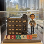 一款為聖類斯中學的黎德維校長（Mr. Lai Tak Wai）訂製的校長退休禮物。精緻的人像公仔手持數學書，莊重地站在一座細節豐富的聖類斯中學立體學校模型旁，底座前方陳列著五個社（LILY, ROSE, SHAMROCK, THISTLE, TULIP）的標誌，整件作品置於透明保護罩中，是一份極具份量的榮休紀念品。