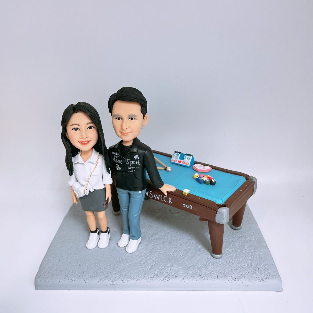 香港訂做情侶桌球公仔，一對情侶公仔企埋一齊，男仔着黑色上衣同藍色褲，女仔着白色恤衫配灰色裙，後面有藍色檯布嘅桌球檯，上面放咗桌球桿、彩球同計分牌裝飾。