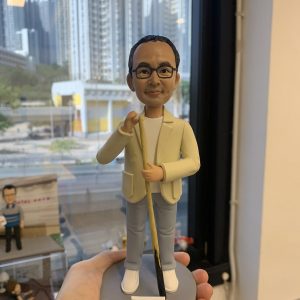 香港訂做生日桌球公仔，男士造型戴黑框眼鏡，着米色外套、白色上衣同藍色長褲，手攞桌球桿，底座有「Joe Ho 生日快樂」字樣，背景為城市高樓景色。