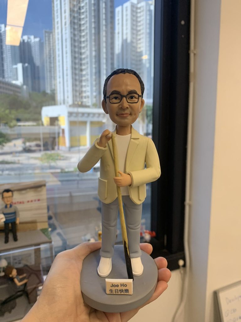 香港訂做生日桌球公仔，男士造型戴黑框眼鏡，着米色外套、白色上衣同藍色長褲，手攞桌球桿，底座有「Joe Ho 生日快樂」字樣，背景為城市高樓景色。