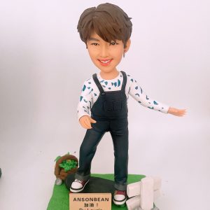 定制滑板公仔，短髮笑容造型，身穿黑色背帶褲與白色印花長袖上衣，站在綠色滑板造型底座上，旁邊有小籃與木牌裝飾，木牌寫有「ANSONBEAN 加油！By Laurie」。