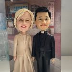 雙人牧師人像公仔並肩站立於淺灰石質底座：右側人物穿黑色西裝搭白色牧領、黑鞋，臉上帶溫和笑容；左側人物身著米色套裝與及膝洋裙、淺金短髮，神情親切。底座前方白色銘牌印有「牧師榮休紀念」；背板是教會門面與紅色雨棚、中文招牌的建築外觀。整體細節精緻、姿態自然，適合作為牧師榮休與教會致謝的人像公仔禮物。