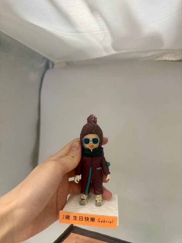 手持底座的小朋友主題人像公仔：戴針織毛帽與墨綠連帽圍脖、穿酒紅色連身外套與運動鞋的bb造型，臉上配藍框圓形墨鏡，站在白色發泡底座上；底座前緣橘色標籤寫有「2歲 生日快樂 Gabriel」字樣；背景為簡潔棚拍白色。