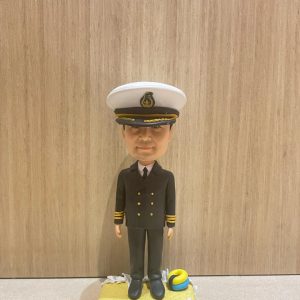 船長主題的人像公仔直立於沙灘質感底座上；戴白色船長帽（黑色帽沿與金色繡飾、帽徽），穿深色雙排扣制服外套與長褲、黑鞋，袖口配金色階級條。底座上點綴貝殼與迷你救生圈等海洋元素。