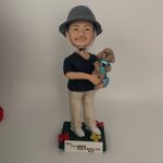 一款站立的人像公仔置於深綠色方形底座上，戴灰色漁夫帽、黑色上衣、米色長褲與白色球鞋；雙手懷抱一隻穿藍綠背帶的棕色捲毛小型犬寵物公仔，犬隻五官圓潤、耳朵下垂，呈現貴賓犬風格。底座四角有紅黃小花裝飾，前方附白色名牌刻字。背景為素白。