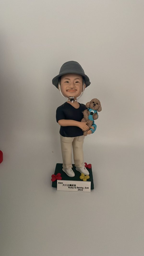 一款站立的人像公仔置於深綠色方形底座上，戴灰色漁夫帽、黑色上衣、米色長褲與白色球鞋；雙手懷抱一隻穿藍綠背帶的棕色捲毛小型犬寵物公仔，犬隻五官圓潤、耳朵下垂，呈現貴賓犬風格。底座四角有紅黃小花裝飾，前方附白色名牌刻字。背景為素白。