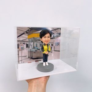 透明盒內的人像公仔站在灰色圓形底座上，身穿黑黃色外套、深色長褲與白鞋，雙手交疊於身前；背板為港鐵車站大堂場景，可見黃黑色資訊牌、客服中心與閘機等設施。整個展示盒由手掌托起，背景為素白牆。