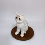 單隻白奶油色相間的坐姿貓公仔置於圓形棕色底座上，粉色鼻頭、黑大眼、耳尖立起；毛流以細緻刻線呈現，鬍鬚纖細可見。背景為簡潔淺灰白色。