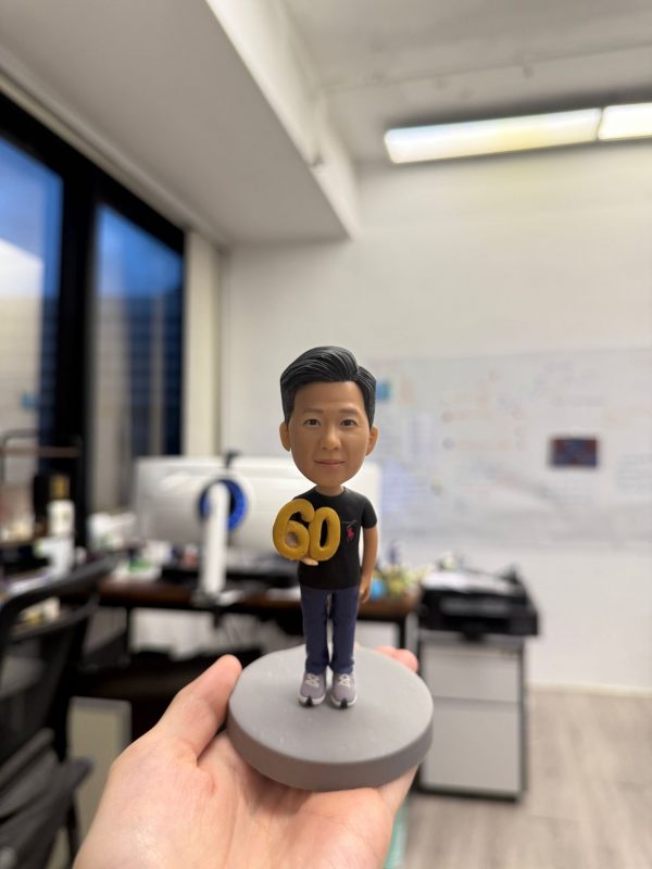 採用站立姿勢，雙手持有一個醒目的金色「60」數字，象徵著重要的里程碑與慶祝時刻。 公仔穿著 黑色休閒上衣 搭配 深色長褲，腳踩 運動鞋，展現簡約隨性的日常風格；髮型整潔俐落，神情帶有微笑，塑造出親切又穩重的形象。 底座以 簡潔圓形灰色設計，讓整體焦點更集中於公仔與「60」的慶祝元素。