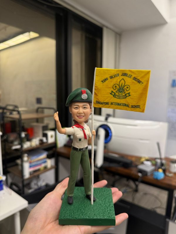 童軍主題人像公仔站在綠色方形底座上，戴綠色貝雷帽與徽章，穿卡其色短袖制服配紅色領巾、綠色長褲與黑鞋，右手高舉、左手持白色旗桿，旗面為黃色並印有童軍百合花徽與英文隊名。