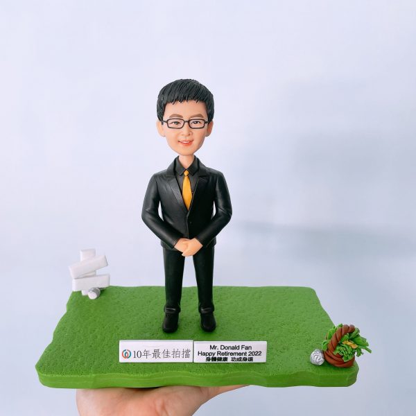 榮休主題的人像公仔：戴眼鏡的短髮男士身穿黑色西裝與金黃色領帶，雙手於身前交握，站在綠色草地質感的地台上；地台前緣設有兩塊金屬銘牌，其一寫有「10年最佳拍檔」，另一塊標示「Mr. Donald Fan Happy Retirement 2022」與祝福字樣。左後方有白色指示牌小道具，右前方擺放以編織圈與綠植構成的小擺飾；整體以淺灰白背景拍攝，手掌托起作品，呈現適合作為高層榮休禮物的人像公仔展示。