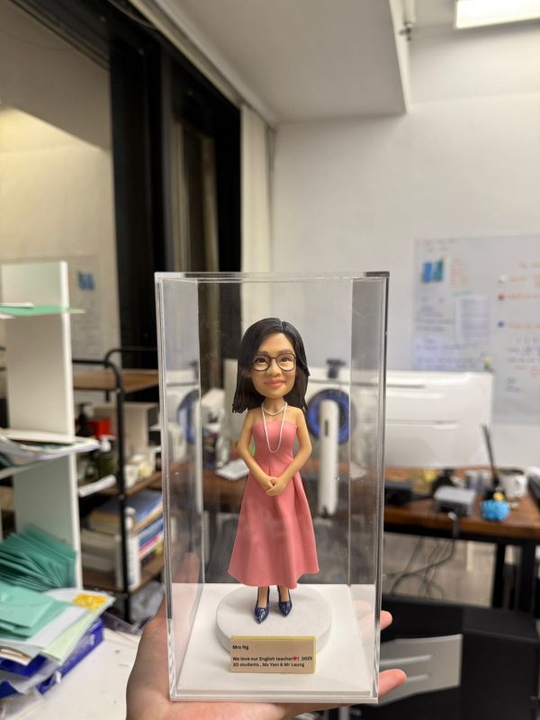 一款人像公仔置於透明壓克力盒內：女性公仔留中長黑髮、戴黑框眼鏡，穿粉色無袖長洋裝與深色高跟鞋，頸上配長項鍊，雙手於身前交握，站在白色圓形底座上。底座前有金色名牌，刻字含「Mrs Ng」「We love our English teacher」「2023」「3D students, Ms Yam & Mr Leung」等英文字樣。背景為辦公室/教職員室環境，可見桌面、文件與架子等雜物。