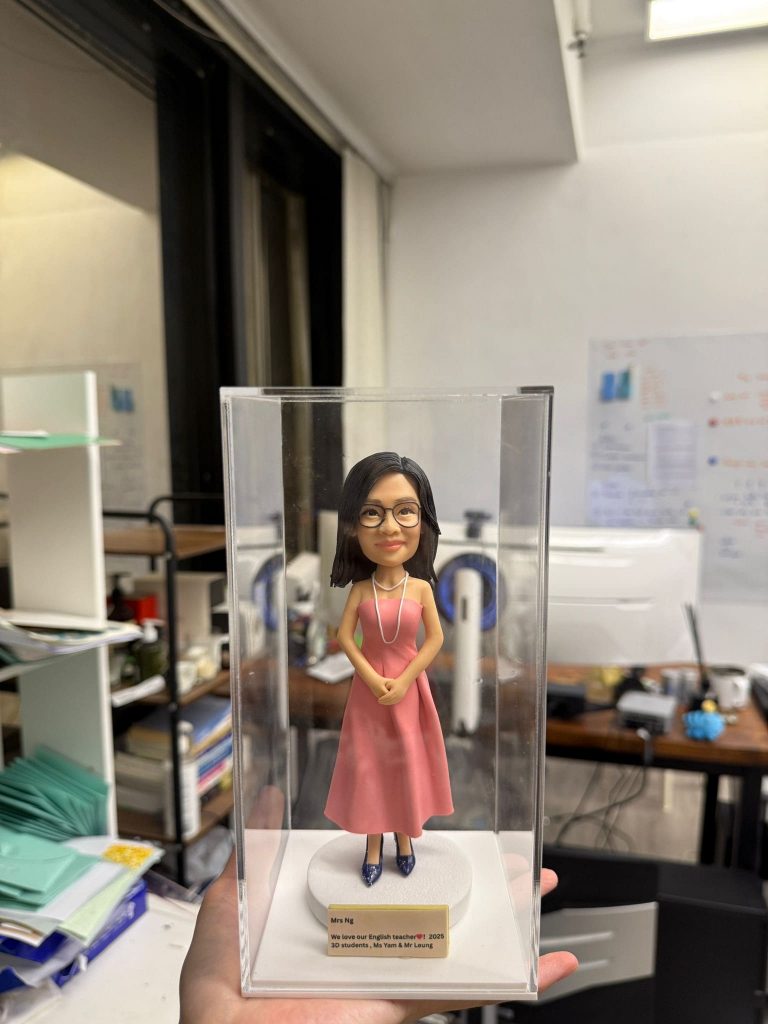 一款人像公仔置於透明壓克力盒內:女性公仔留中長黑髮、戴黑框眼鏡,穿粉色無袖長洋裝與深色高跟鞋,頸上配長項鍊,雙手於身前交握,站在白色圓形底座上。底座前有金色名牌,刻字含「Mrs Ng」「We love our English teacher」「2023」「3D students, Ms Yam & Mr Leung」等英文字樣。背景為辦公室/教職員室環境,可見桌面、文件與架子等雜物。