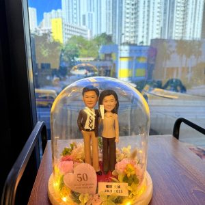 一組長輩主題的人像公仔置於玻璃盅內展示：木質圓形底座上鋪紅色地面與綠植苔面，周圍環繞粉、奶白與淡黃系的玫瑰與葉材。兩位角色並肩站立：左側穿深色外套、白襯衫與棕色長褲，右側穿米色上衣與深色褲，皆著深色鞋。玻璃盅內側繞有暖白燈串，底座前方有粉色愛心立牌寫「50 GOLD WEDDING ANNIVERSARY」，旁邊金色小牌標示日期「28.9.2025」與中文字樣。作品置於木桌上，背後可見窗外高樓城市景與車流。