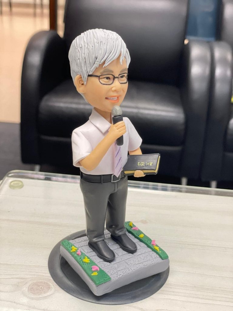 宗教主題的人像公仔：銀灰短髮、戴眼鏡的男性站姿，穿白襯衫配淡紫色領帶與深灰長褲，右手握麥克風，左手持黑色金邊聖經樣小冊；腳踏石磚紋台座，四周綠邊點綴小花。適合作為牧師禮物的人像公仔。