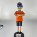 休閒風格的人像公仔雙臂交叉站立於黑色圓形底座上，戴反戴灰色帽、配大型反光墨鏡，穿橘色短袖上衣、深色及膝短褲、藍白襪與黑紅運動鞋；底座前貼有小牌寫著「Dear Wa Happy Birthday Sep 2021」。