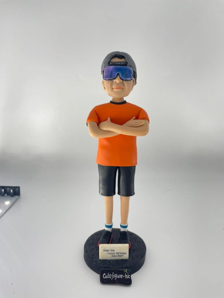 休閒風格的人像公仔雙臂交叉站立於黑色圓形底座上,戴反戴灰色帽、配大型反光墨鏡,穿橘色短袖上衣、深色及膝短褲、藍白襪與黑紅運動鞋;底座前貼有小牌寫著「Dear Wa Happy Birthday Sep 2021」。