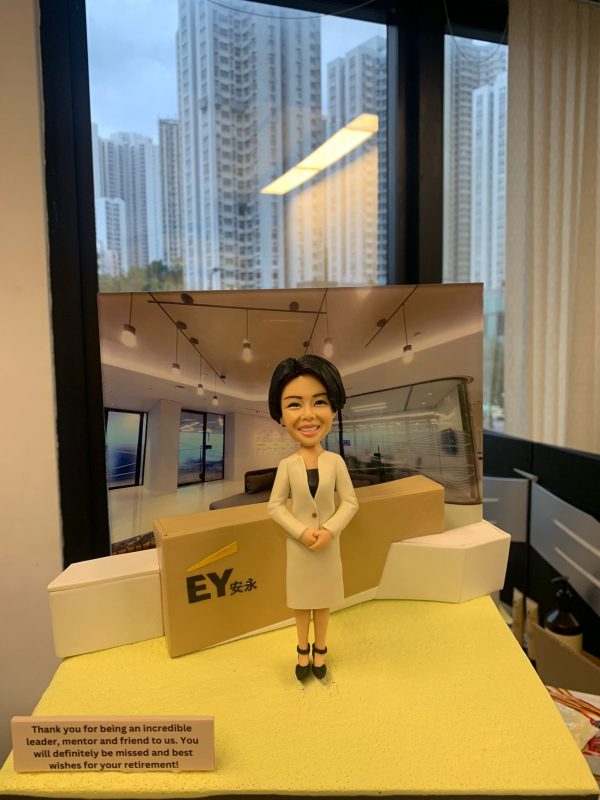 四大會計師所主題的人像公仔：短髮女性穿米色套裝外套與黑色內搭，雙手於身前交握，站在帶有 EY安永 標誌的接待台前；背板呈現現代化辦公室與天花吊燈設計，台前左側有英文字樣的榮休感謝銘牌。場景放置在窗景前的桌面上，整體營造出專業、親切的事務所接待區氛圍，適合作為 EY安永 高層榮休禮物與會計師人像公仔展示。