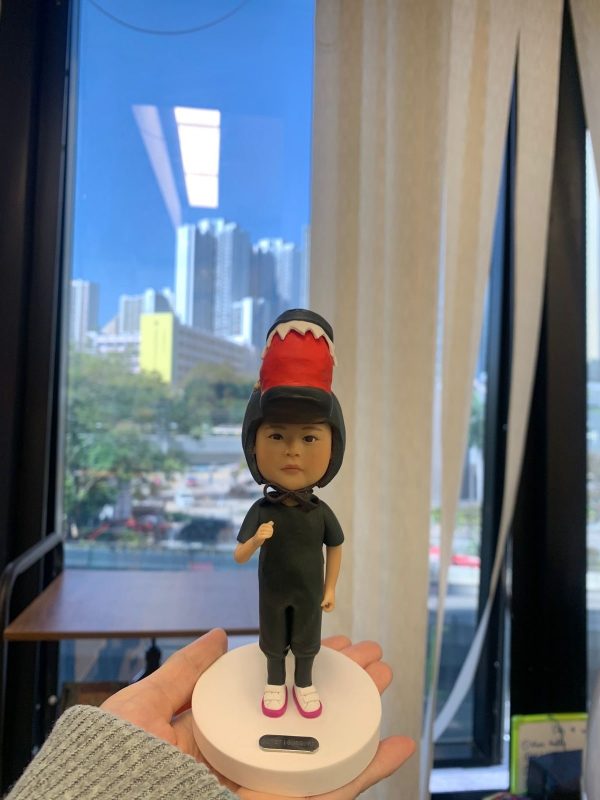 一款小朋友／bb主題的人像公仔立於白色圓形底座上，被手掌托著展示。公仔穿全黑連身服與黑色安全帽，帽頂裝飾紅色帶白齒狀邊的恐龍頭造型；腳穿白底粉邊鞋，胸前綁有黑色蝴蝶結。背景是大窗外的城市高樓與藍天，室內有米色窗簾與木桌。