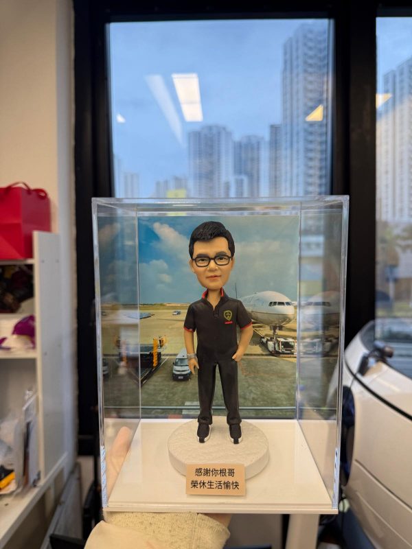 男性人像公仔置於透明展示盒內，站在白色圓形底座上；公仔戴黑框眼鏡，短髮，穿深色短袖工作POLO衫與長褲、黑鞋，胸前有盾形徽章與小國旗樣式貼章，一手插袋一手自然下垂。底座前方貼有小牌寫著「感謝你根哥 退休生活愉快」。