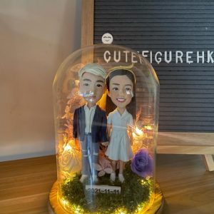 木質底座上的玻璃永生花燈罩作品：內含一對情侶人像公仔，左側角色著深藍外套與破洞牛仔褲、白鞋；右側角色穿白色V領短袖連身裙與白鞋。腳下覆苔綠草，周圍點綴紫玫瑰與乾燥花材，環繞暖白色小燈串；前方小牌寫有日期「2021-11-11」。背景為木桌與置有字母板的場景。