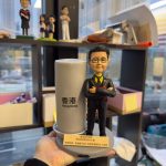 手掌托著一座港鐵主題人像公仔擺設：公仔穿黑色西裝配黃黑襯衣與領帶、雙手交叉於胸前，站在灰色圓形底座上；旁側設有圓柱形站牌模型，印有「香港 Hong Kong」字樣；底座前方貼有金色感謝字牌。背景為室內層架與窗景，可見其他公仔模型與城市高樓。