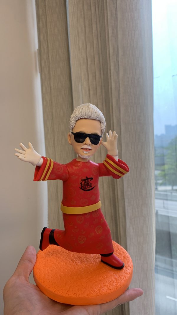 戴黑色墨鏡、白色短髮與小鬍子的男性人像公仔，單膝前跨、雙臂張開的動感姿勢；身穿紅色中式長袍，衣身帶有低飽和暗花紋理，胸前為黑色書法風圖案，袖口三道金色條紋，腰間繫金色腰帶，腳穿紅黑靴。公仔站在橘色多孔質感的圓形底座上；背景為室內窗簾與窗外街景，有人手掌托住底座。