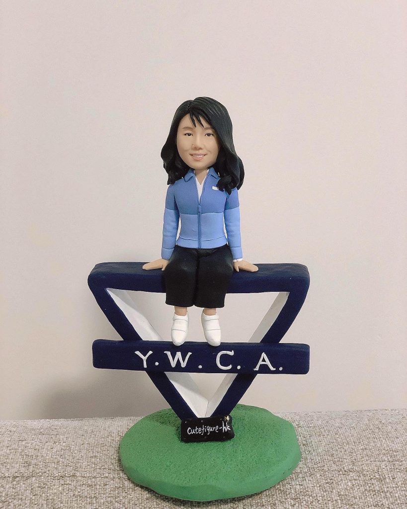 女同事主題的人像公仔坐在深藍色「Y.W.C.A.」字樣的三角造型標誌上；公仔長髮、微笑，穿淺藍外套與深色半身裙、白色鞋子。標誌立於綠色草地質感圓形底座上，前方有小黑牌寫著「cutefigure-hk」。