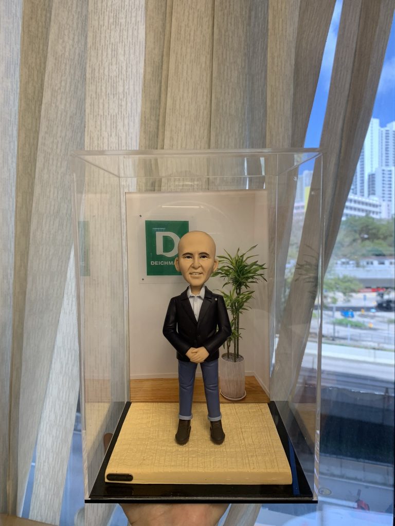 辦公室主題的人像公仔：一位外國男性老闆形象，光頭、微笑表情，穿黑色西裝外套與白襯衫、藍色反摺長褲與棕色短靴，雙手於腹前相合站姿。公仔置於木紋地台，後方有綠植與牆上綠白色公司標誌牌，整體收納在透明防塵展示盒中；背景為落地窗簾與城市景觀。作品呈現適合作為老闆或上司的祝福禮物的人像公仔。