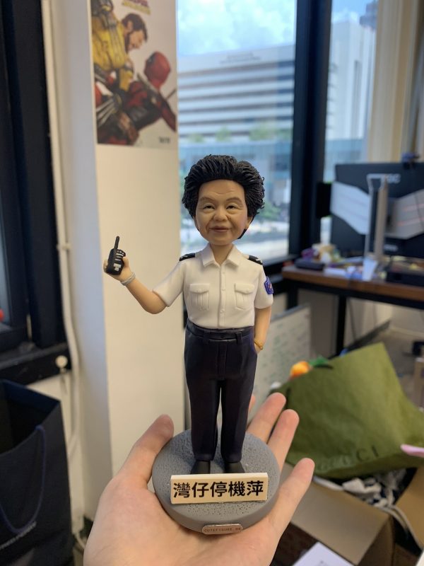 一款女性人像公仔站在灰色圓形底座上，底座前有白底黑字的牌子；公仔右手持對講機、左手自然垂放，身穿白色短袖制服襯衫（有肩章與口袋）與深色長褲、黑色皮鞋，袖臂見圓形臂章。表情親切微笑，短卷髮造型。背景為室內辦公環境，有窗外城市建築、桌面與紙箱雜物可見；一隻手掌托住底座。