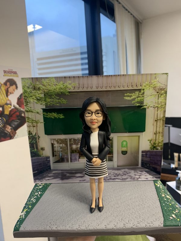 黑長髮戴眼鏡的女性人像公仔，身穿黑色西裝外套與白色上衣，搭配黑白橫條紋窄裙與黑色高跟鞋；雙手於身前交握、端正站立並微笑。公仔置於灰色粗紋質感底座，兩側有綠色點綴，背板為綠色遮陽棚與玻璃門的街景店面，周圍有綠植與牆面細節。