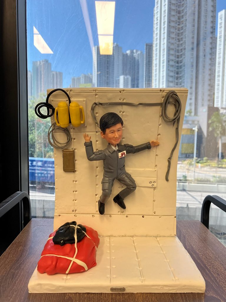 一座情景式人像公仔：男性角色穿灰色連身工作服，胸前貼有「JPL」字樣貼章，雙臂外伸、雙腳彎曲，呈失重漂浮姿態，固定在白色鉚釘結構艙壁前。艙壁左上方掛有黃色裝置與黑色電線，旁邊繞著灰色管線；右側亦有環狀纜線。地面左前方放置以白色綁帶固定的紅黑色軟包裝備。整體置於木桌上，背景是窗外的高樓城市景色與藍天。