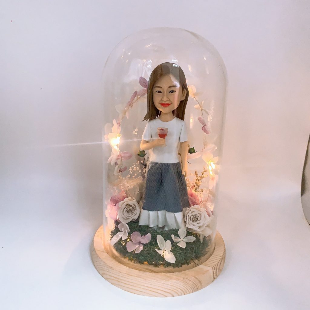 玻璃罩永生花擺設中的女性人像公仔：棕色長髮、微笑神情，穿白色短袖上衣與灰藍色長裙，右手捧著小朵玫瑰。木質底座上鋪設苔綠草地與白、粉、淺紫色永生花與花瓣點綴，環繞暖黃小燈串形成花環效果，整體呈現溫柔浪漫、可長久保存的紀念氛圍。