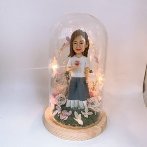 玻璃罩永生花擺設中的女性人像公仔：棕色長髮、微笑神情，穿白色短袖上衣與灰藍色長裙，右手捧著小朵玫瑰。木質底座上鋪設苔綠草地與白、粉、淺紫色永生花與花瓣點綴，環繞暖黃小燈串形成花環效果，整體呈現溫柔浪漫、可長久保存的紀念氛圍。