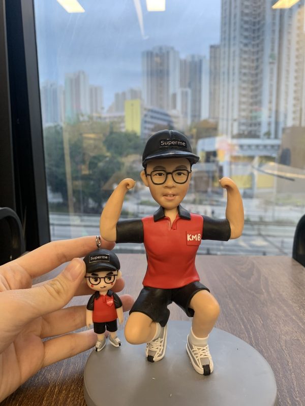 一組人像公仔擺設放在木桌上：主體公仔戴黑色鴨舌帽（上有「Superme」字樣）、黑框眼鏡，穿紅黑配色短袖上衣左胸印有「KMB」字樣、黑色短褲與白色運動鞋；姿勢為單膝下蹲並雙臂彎曲展示肌肉。旁邊手指捏著一個迷你版吊飾公仔，服飾與帽款同款。兩者共同置於灰色圓形底座，背景是玻璃窗外的高樓城市景觀。