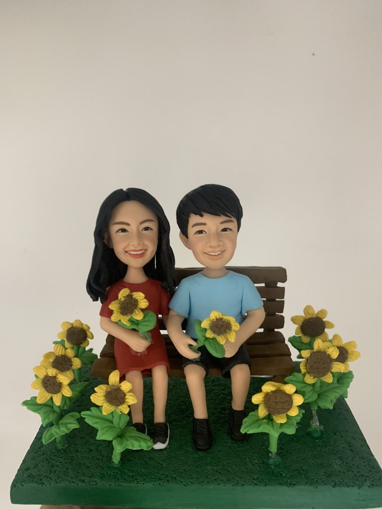 一組情侶人像公仔坐在木質長椅上，置於深綠草地質感底座中央；左側角色穿紅色連衣裙、手抱一束太陽花，右側角色穿淺藍上衣與黑色短褲、手持一朵太陽花。草地兩側與前方環繞多株盛開的太陽花，花心棕色、花瓣亮黃，整體呈現花田氛圍。